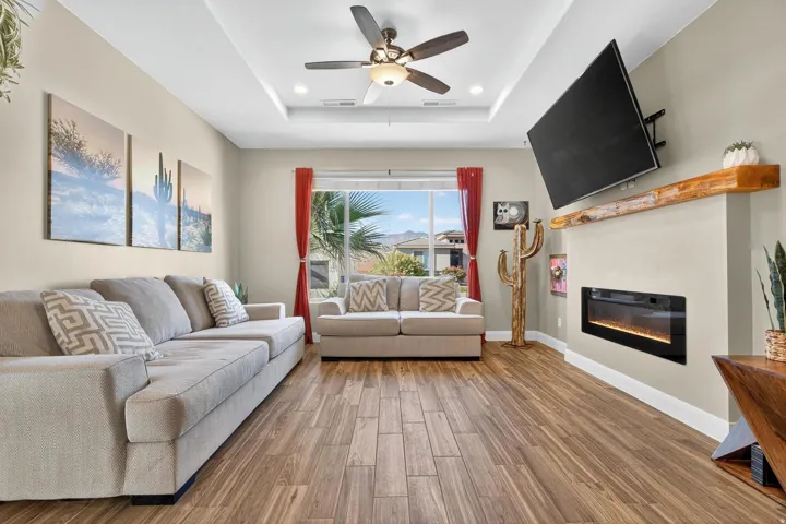 Living area featuring wood-finish flooring, a tray ceiling with a ceiling fan, a built-in electric fireplace with a wood mantel, and large windows