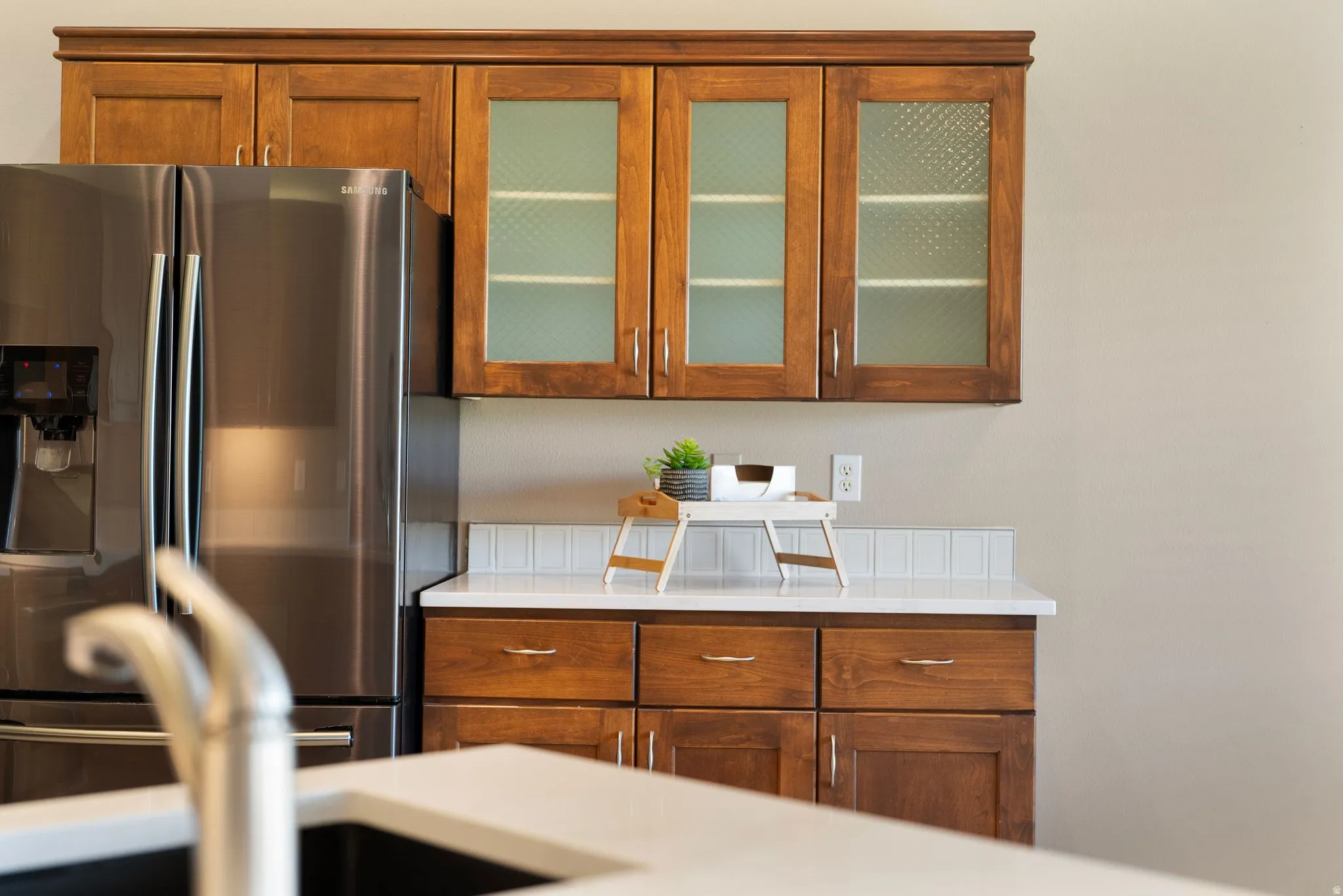 Kitchen featuring wood cabinetry with glass-front uppers, stainless steel refrigerator, white subway tile backsplash, and light-toned countertops