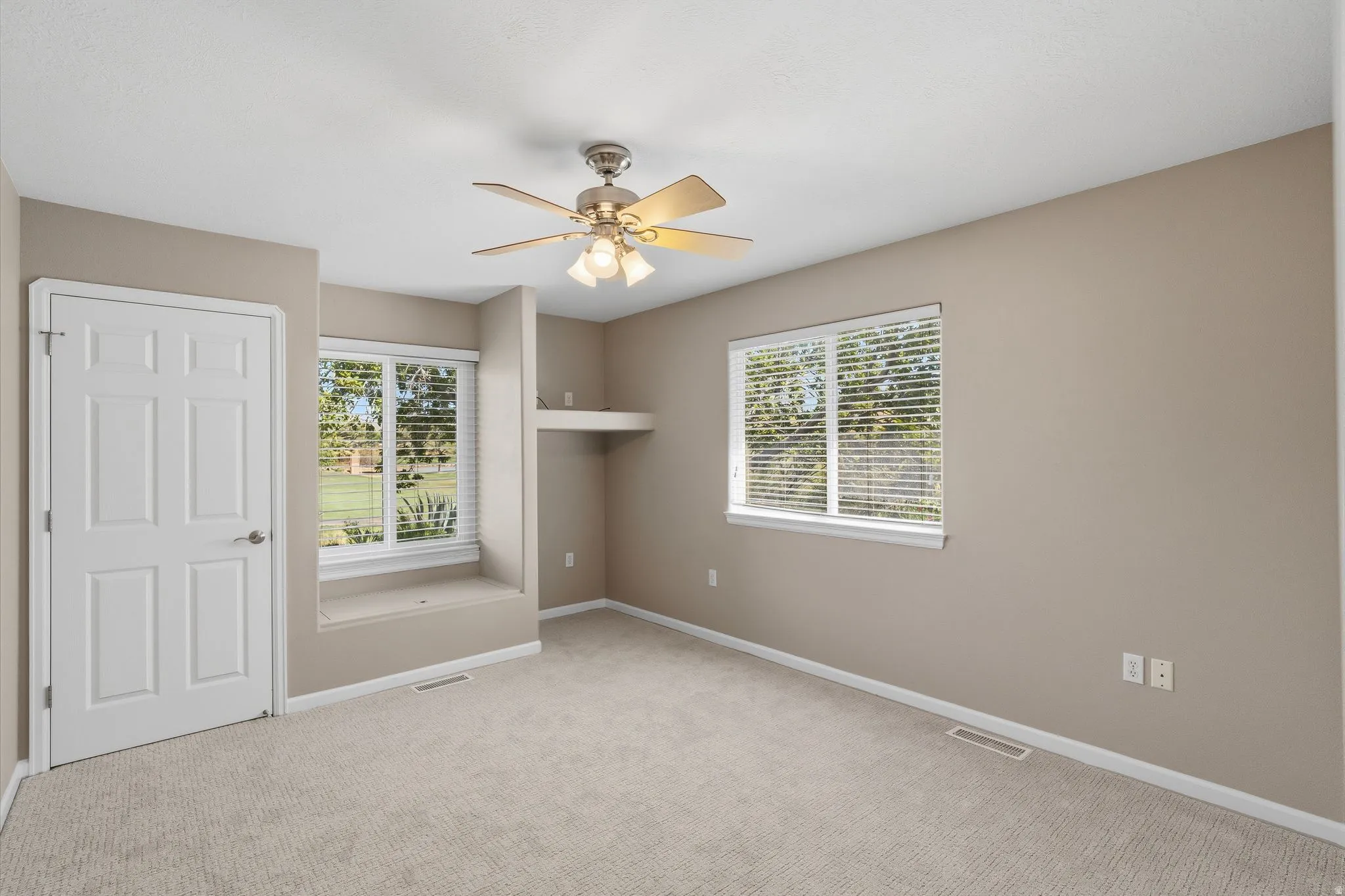 Carpeted room featuring neutral wall tones, two large windows with blinds, a built-in window bench, a ceiling fan with light fixture, and a recessed shelving area