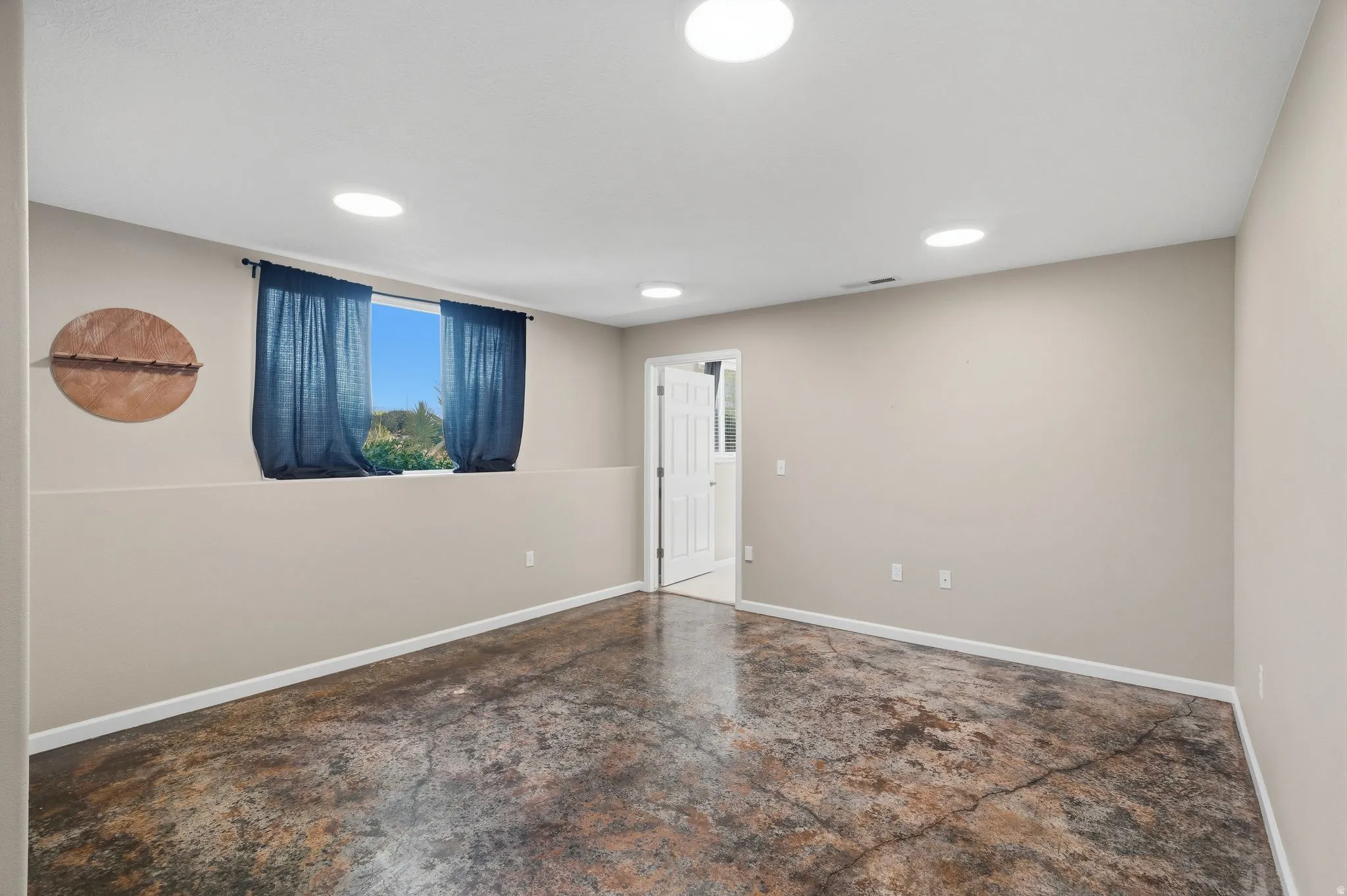 Spacious room featuring a window with natural light, recessed lighting, neutral wall tones, white trim, and a unique stained concrete floor