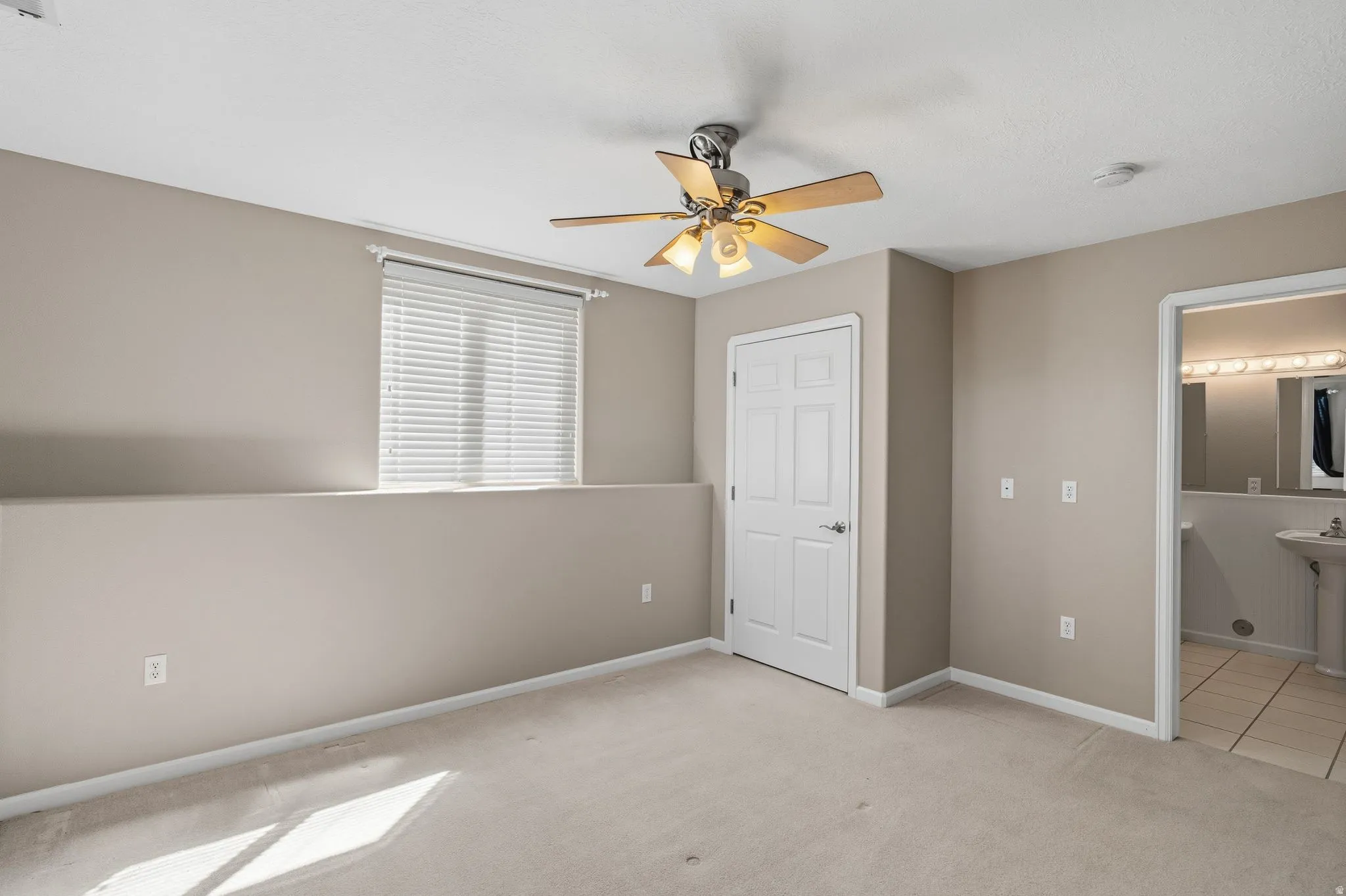 Carpeted interior space featuring a ceiling fan with light kit, a window with horizontal blinds, a six-panel door, and access to a bathroom with tile flooring and a pedestal sink