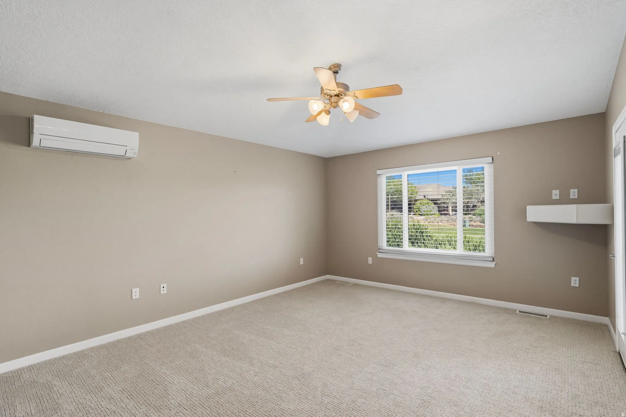 Carpeted interior space featuring a ceiling fan, a wall-mounted air conditioning unit, a large window with blinds, and a white floating shelf