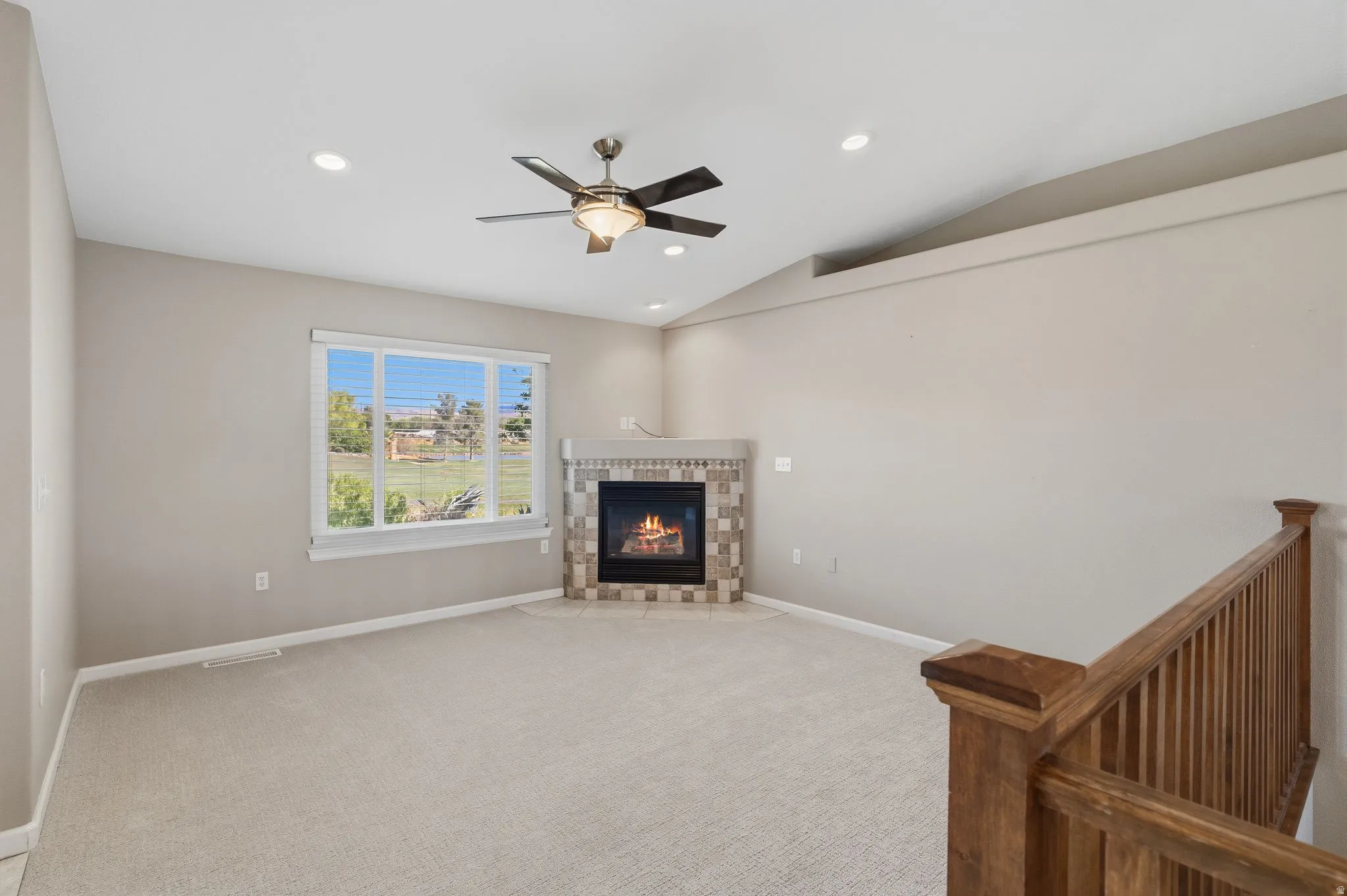 Carpeted living area featuring a corner fireplace with stone tile surround, a large window, a ceiling fan, and recessed lighting