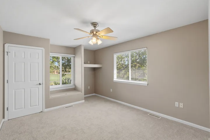 Carpeted room featuring neutral wall tones, two large windows with blinds, a built-in window bench, a ceiling fan with light fixture, and a recessed shelving area
