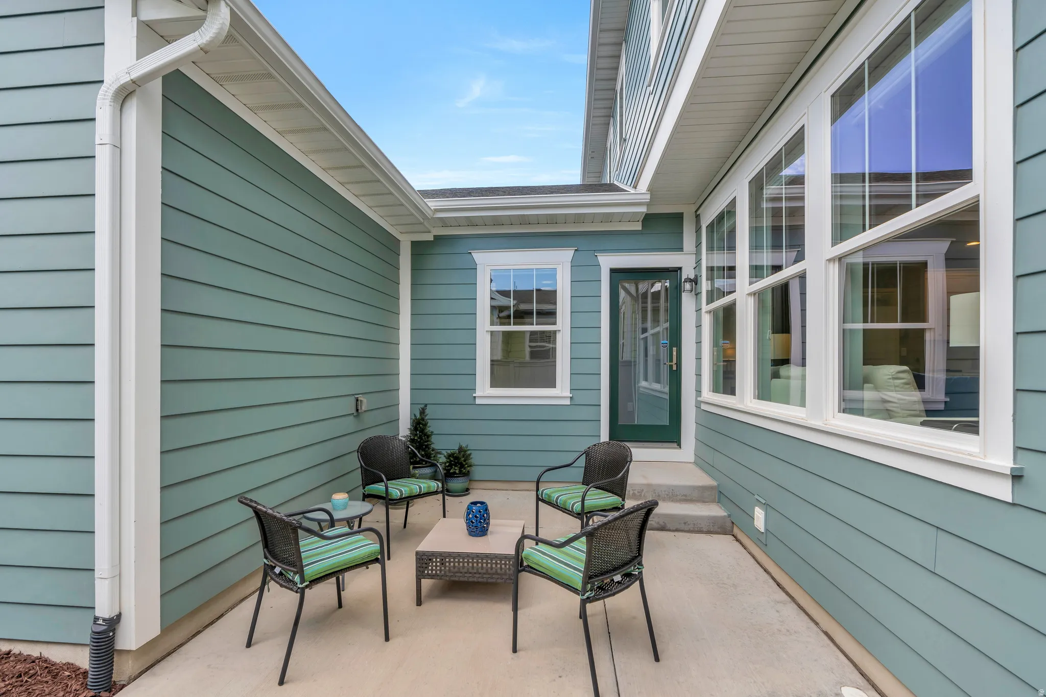 Concrete patio area featuring light blue horizontal siding, white trim, a dark green glass-paneled door, and large multi-pane windows