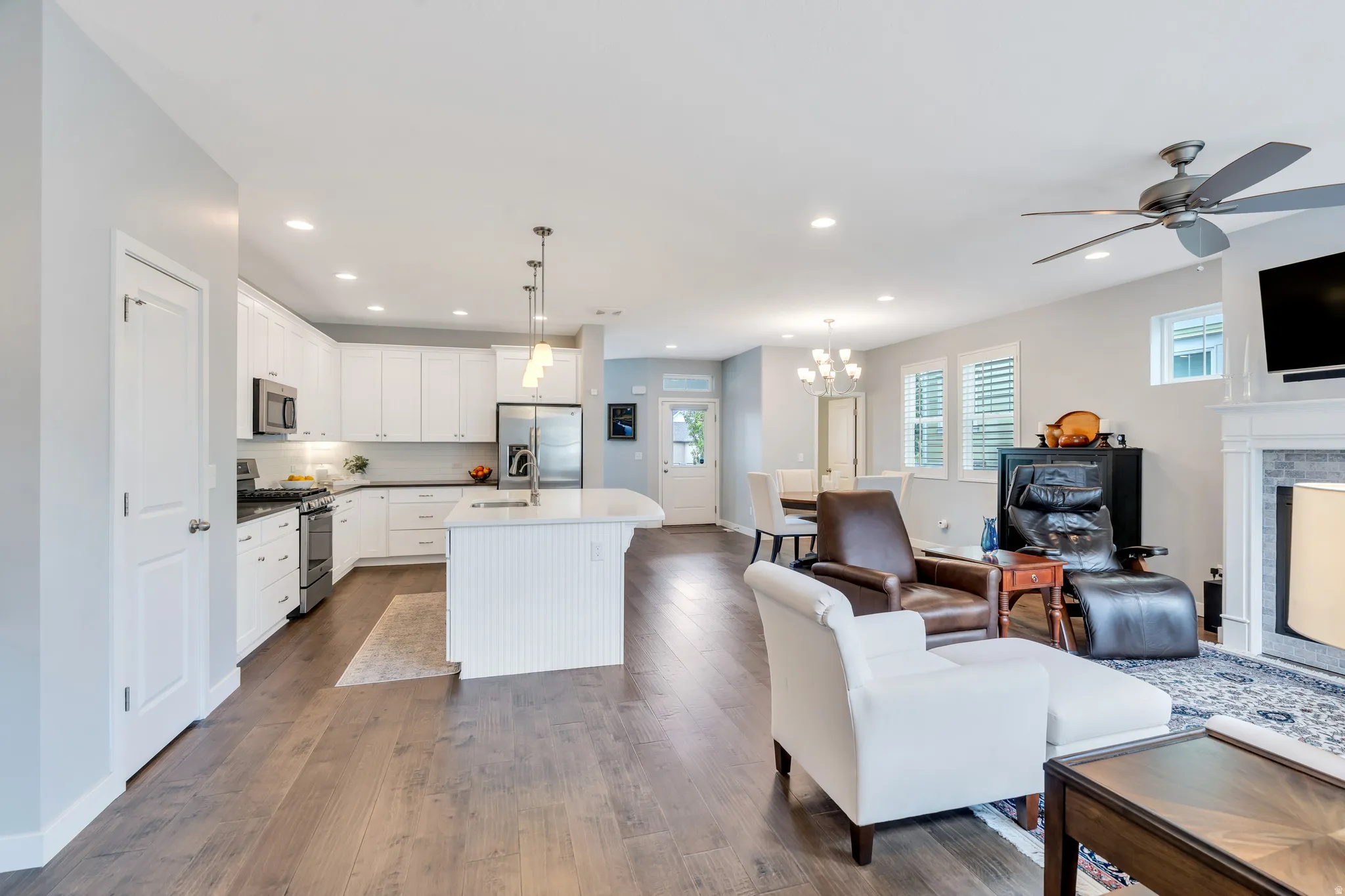 Open-concept living space featuring wood-finish flooring, a kitchen with white cabinetry and an island, a dining area with a chandelier, and a living area with a fireplace and ceiling fan