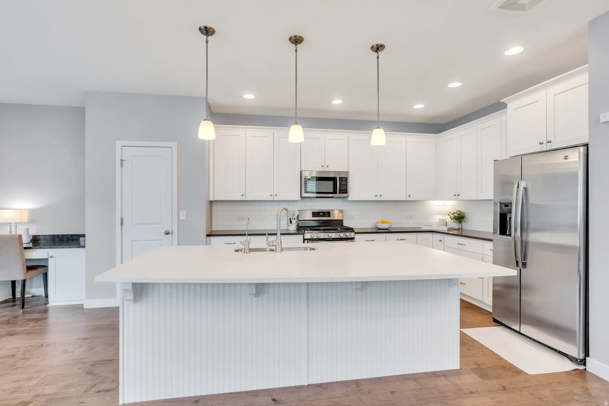Open-concept kitchen featuring a large central island with pendant lighting, white shaker cabinetry, stainless steel appliances, and dark countertops