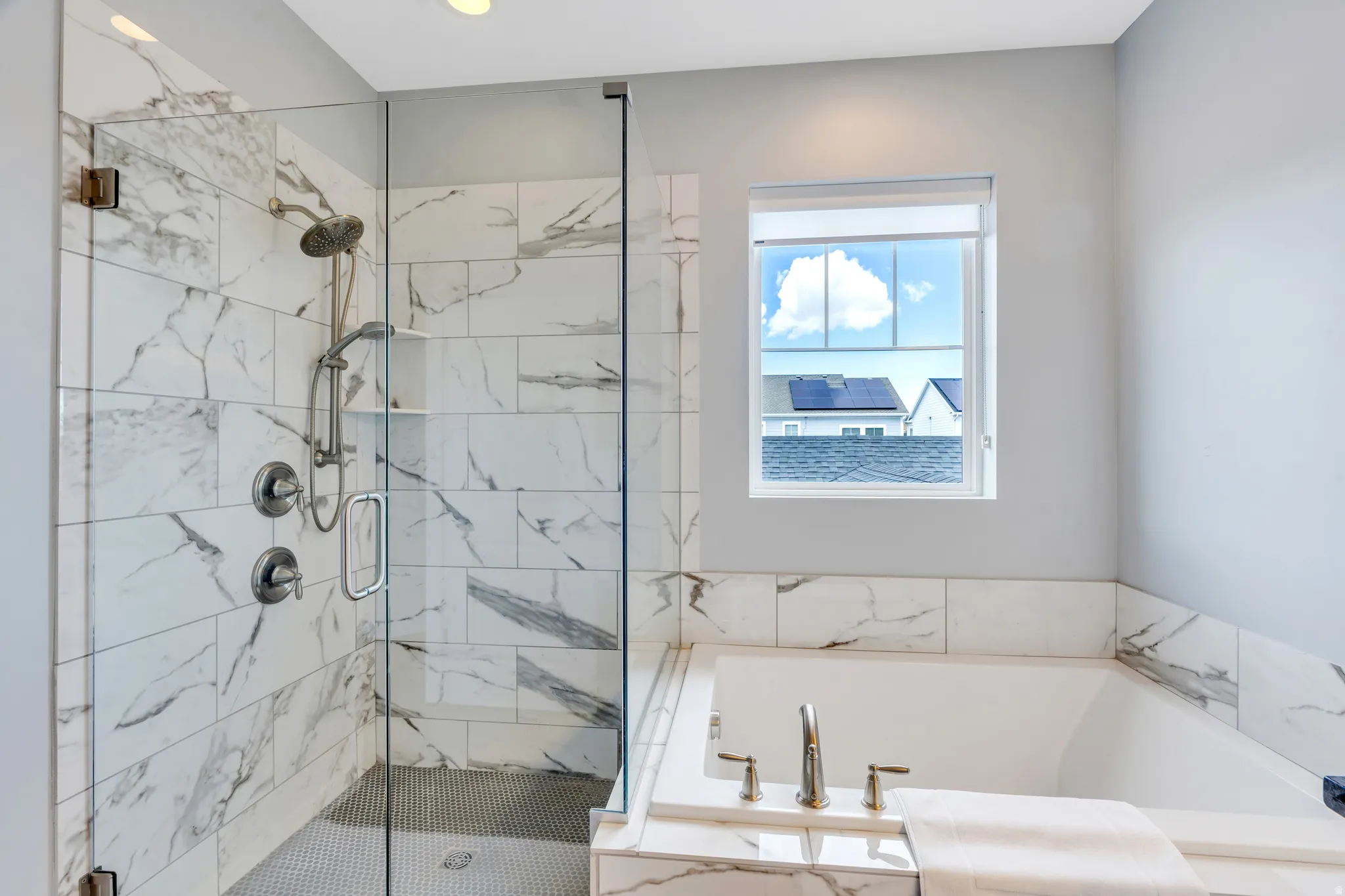 Spacious bathroom featuring a glass-enclosed shower with white and grey tile surround, a built-in shower niche, a soaker tub with matching tile surround, and a single window