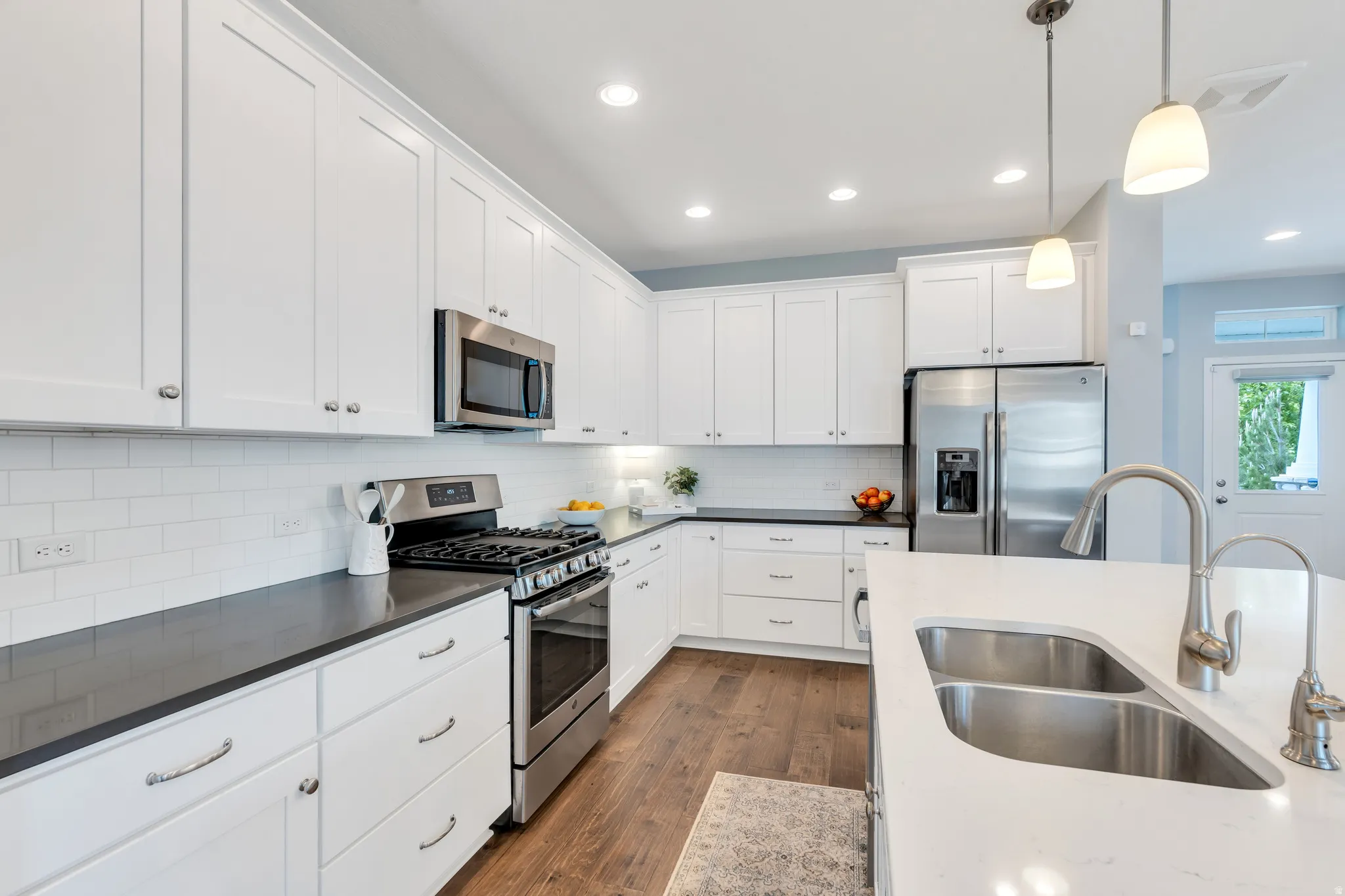 Bright kitchen featuring white shaker cabinetry, stainless steel appliances, dark countertops, a subway tile backsplash, and wood-finish flooring