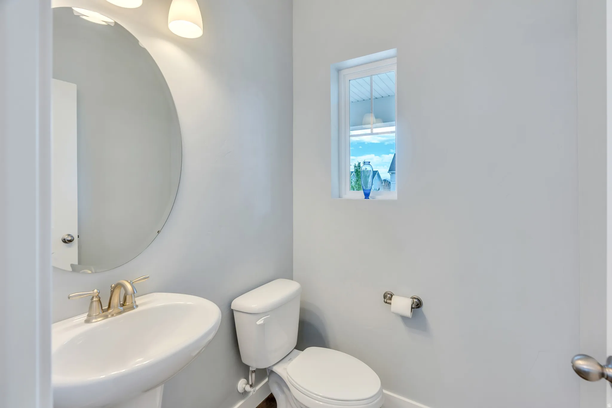 White pedestal sink with a gold-tone faucet, accompanied by a round wall-mounted mirror and a three-light vanity fixture