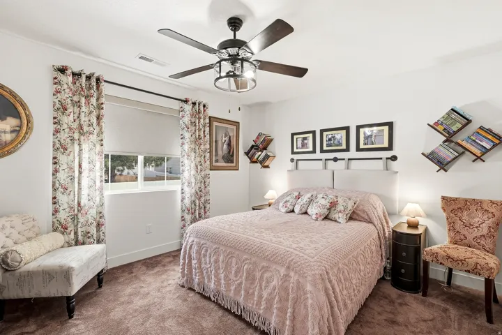 Spacious bedroom featuring light-toned walls, a ceiling fan with integrated lighting, a window with roller shades, and plush carpeting