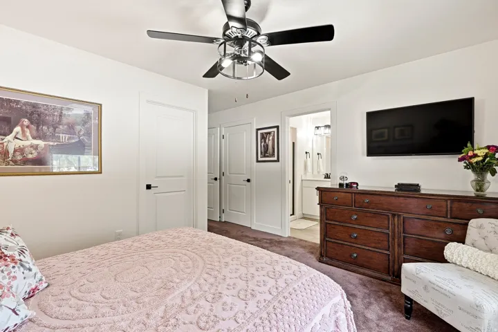 Carpeted room featuring a ceiling fan with light fixture, white interior doors with black hardware, and a partial view of a bathroom with a dual vanity and contemporary light fixture
