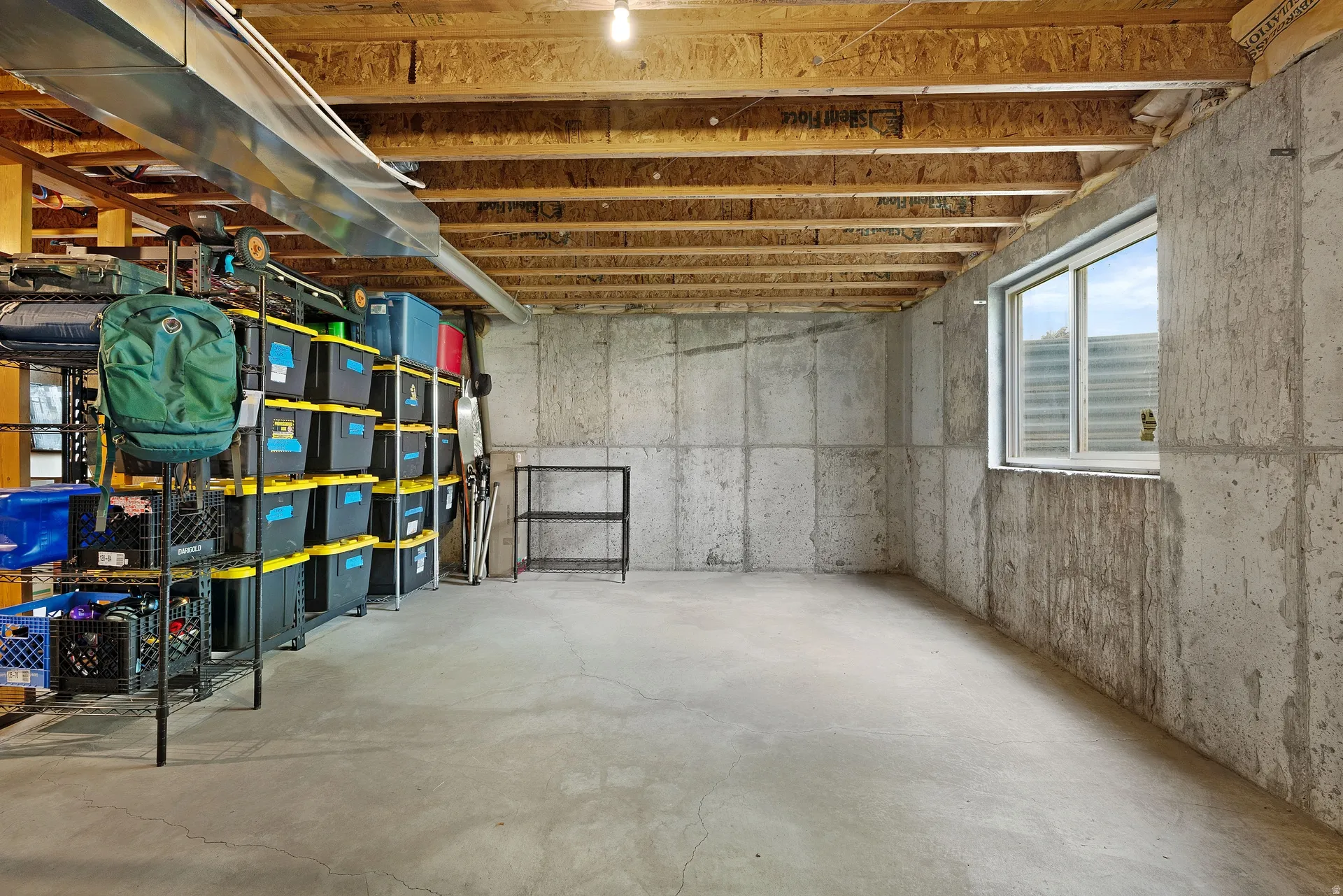 Unfinished basement featuring concrete walls and flooring, exposed ceiling joists, and a horizontal sliding window