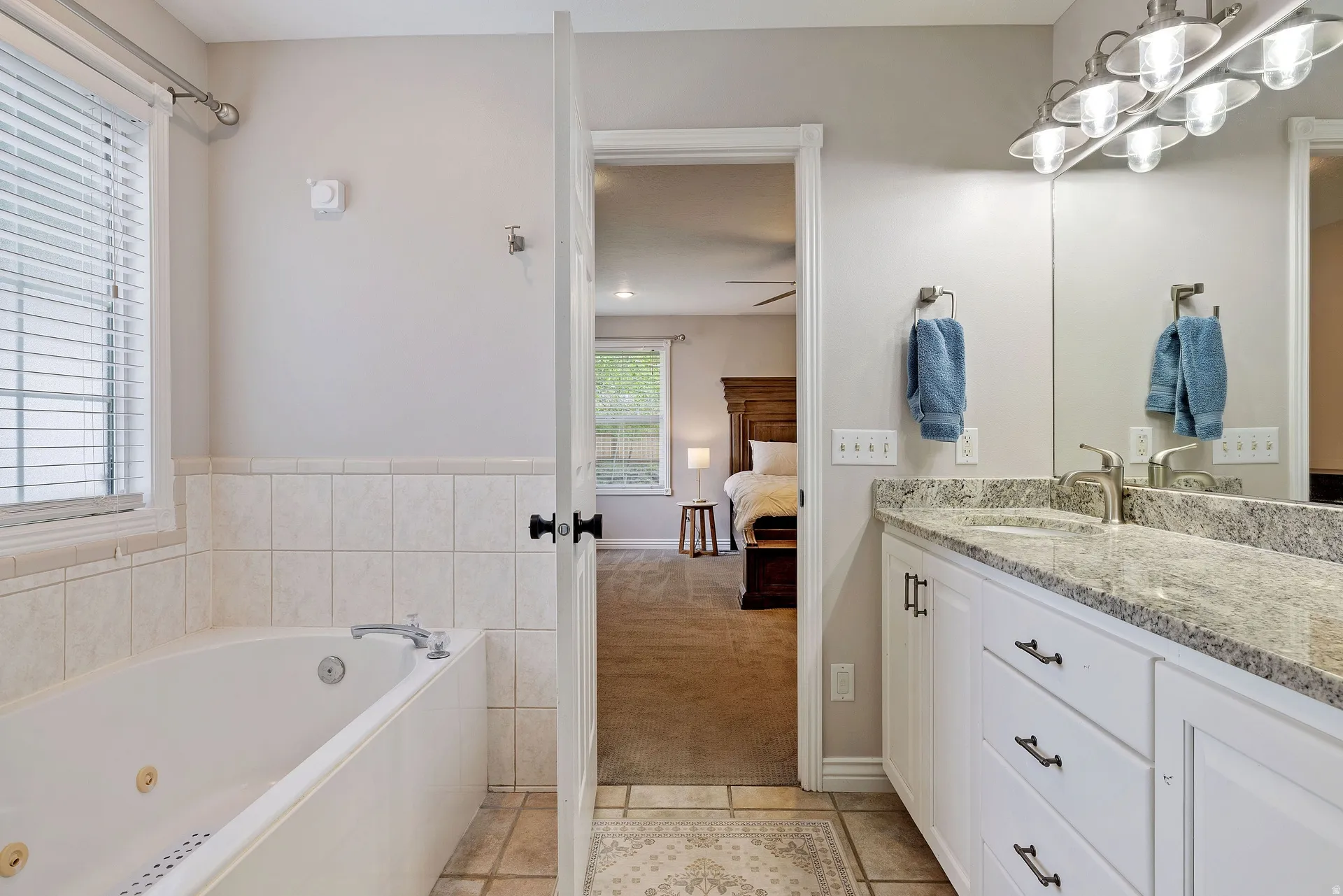 Bathroom featuring a built-in jetted tub, a window with horizontal blinds, tiled flooring, a double vanity with a light-colored granite countertop, and a large mirror with overhead lighting