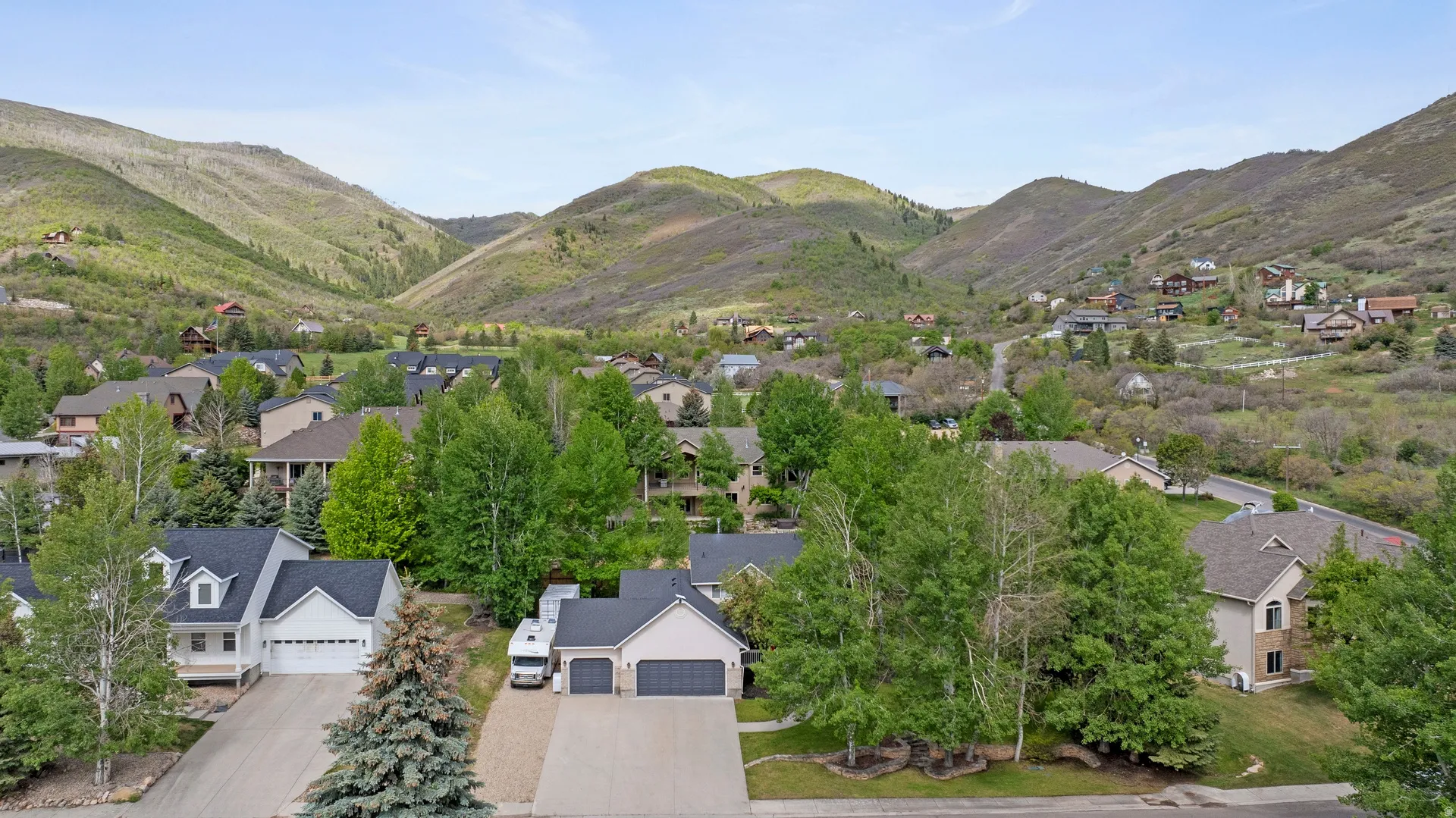 Mountainous terrain surrounds a residential area featuring multiple homes with varied architectural styles, mature trees, and paved driveways
