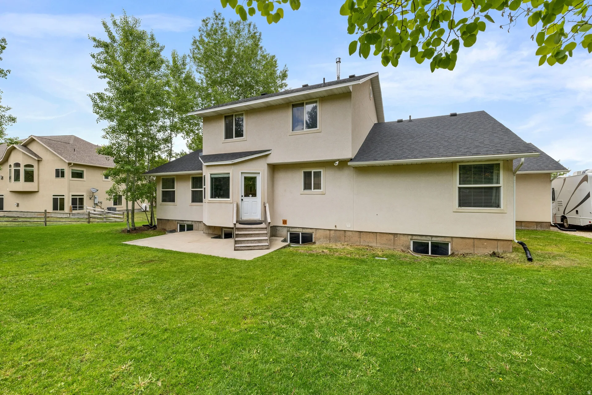 Expansive lawn with a concrete patio, exterior access via a rear door with wood steps, multiple windows, and mature trees providing natural screening