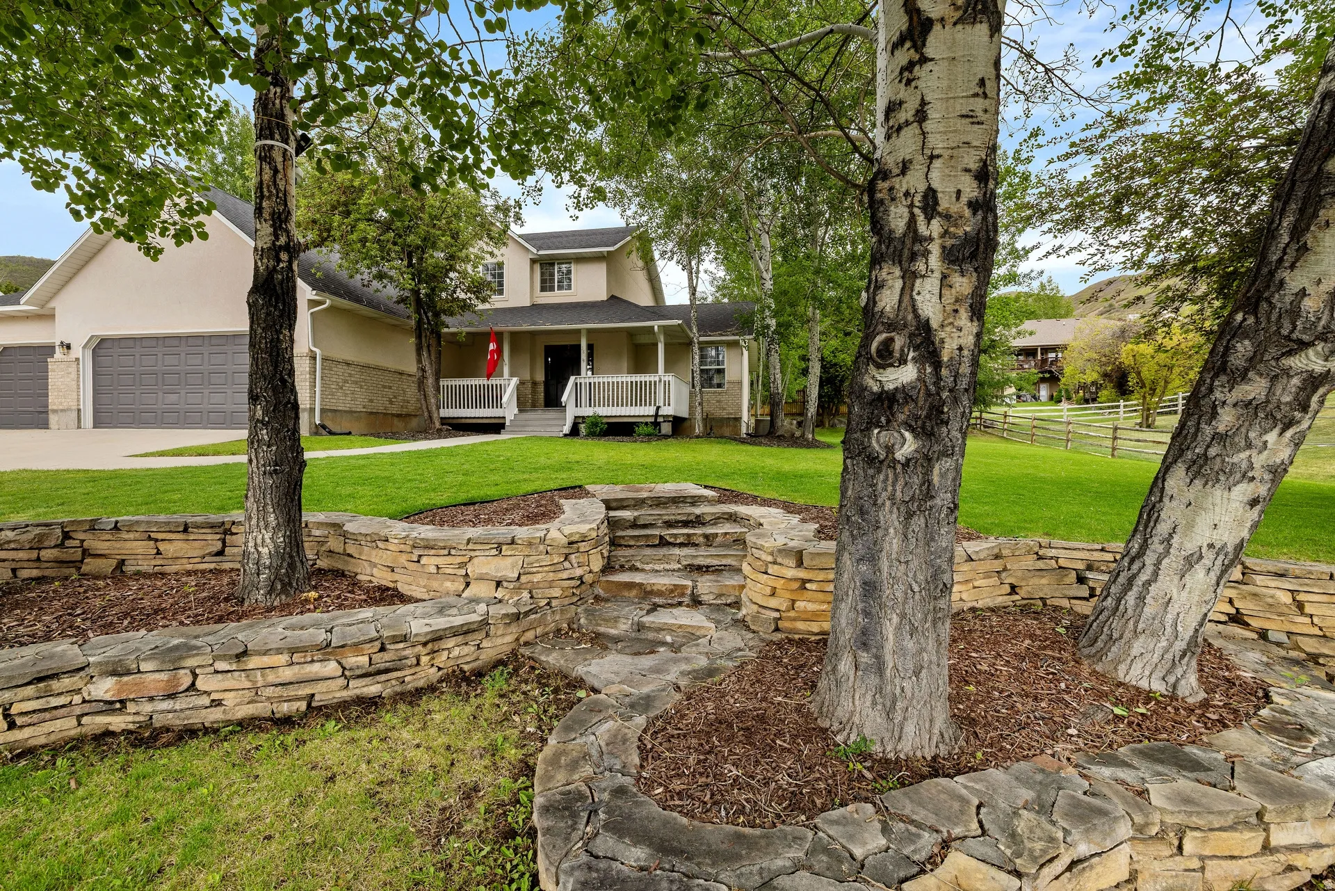 Stone retaining walls and steps lead to a manicured lawn and a stucco exterior with a covered porch and white railing