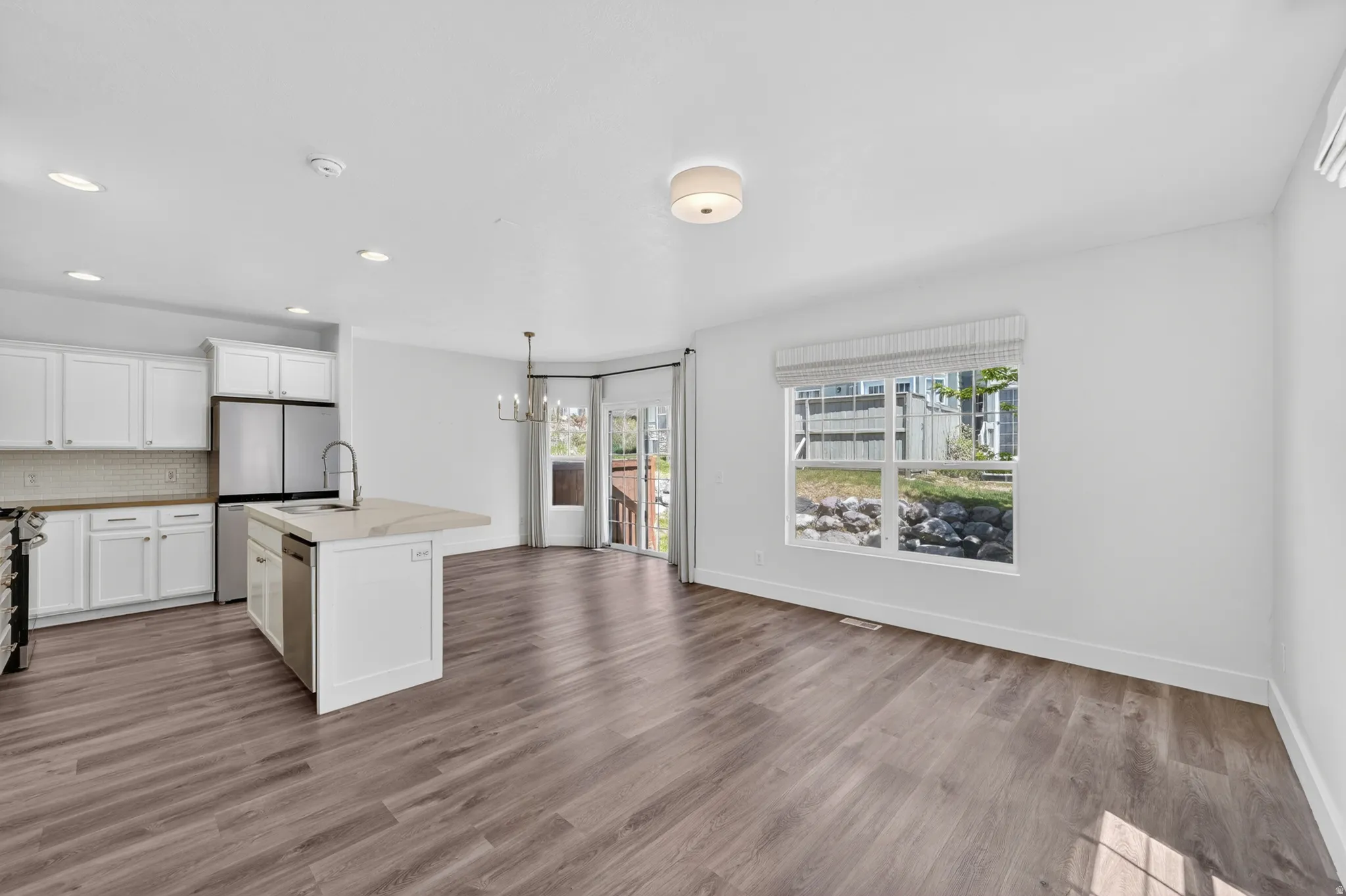 Open-concept living area featuring wood-finish flooring, a kitchen island with sink and integrated dishwasher, white cabinetry with stainless steel appliances, and a dining area with a chandelier