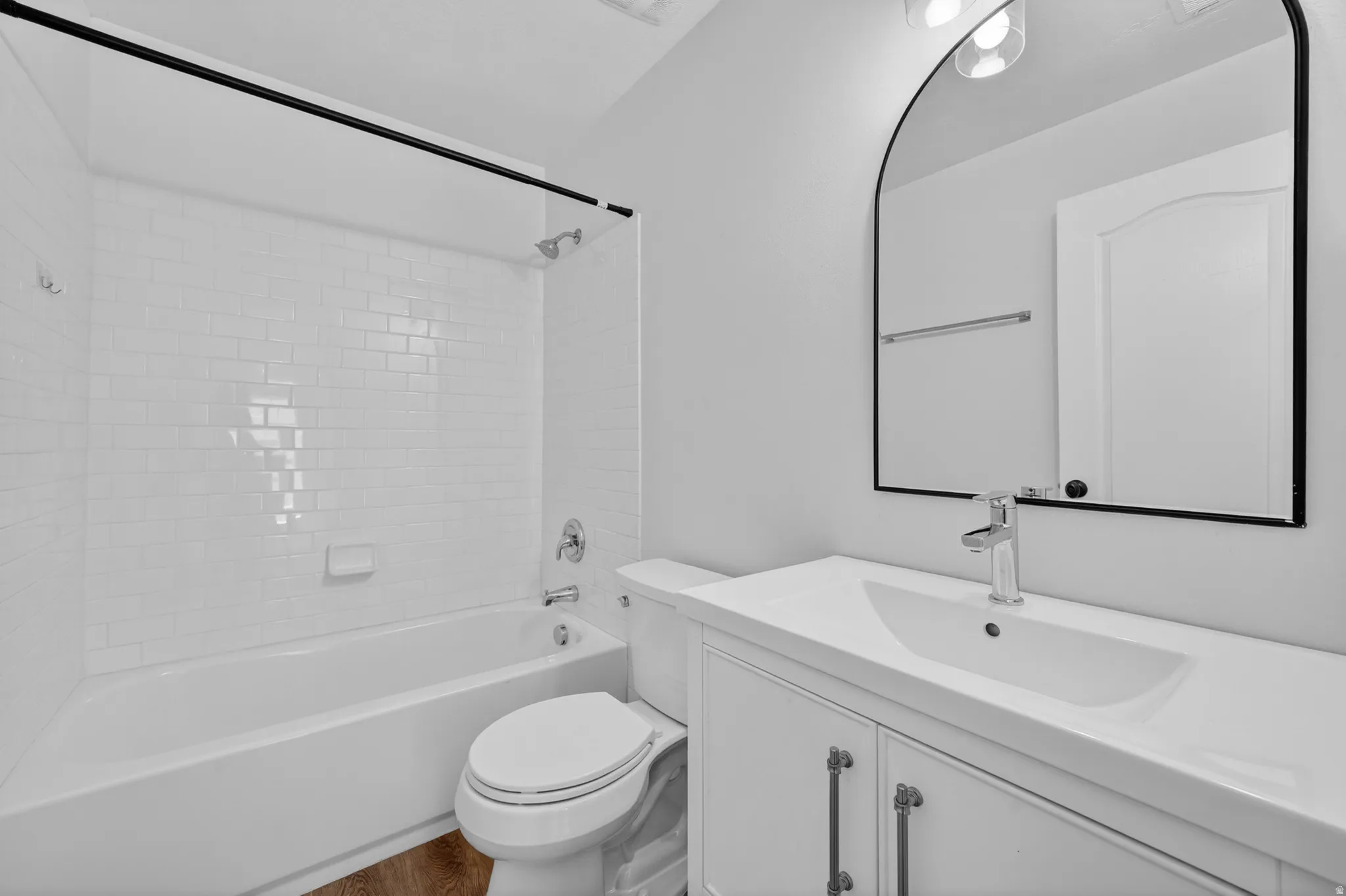 Bathroom featuring a white subway tile tub surround, white vanity with integrated sink, chrome faucet, arched mirror, and wood-finish flooring