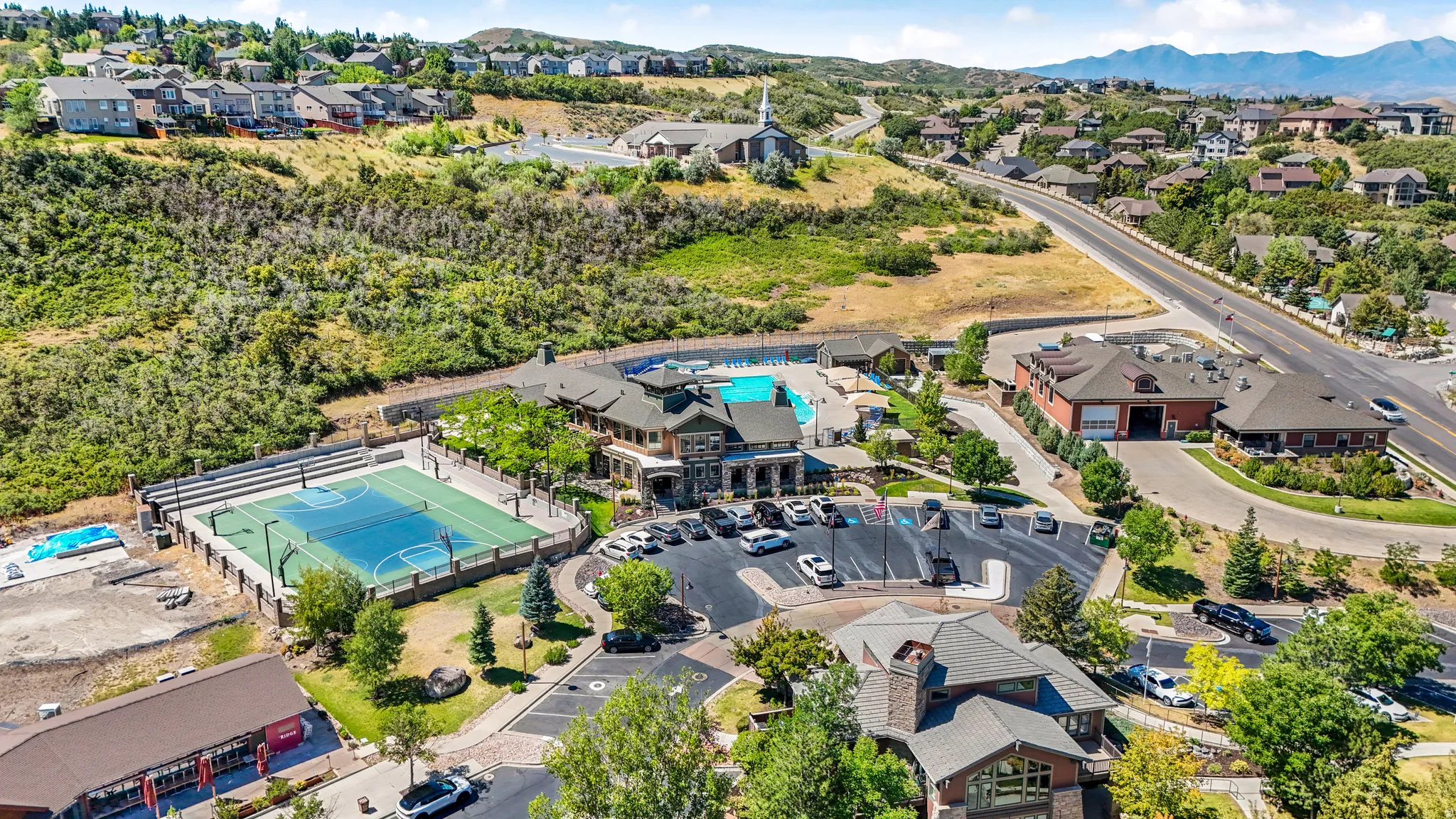 Expansive recreation area featuring a multi-sport court with spectator seating, a large outdoor swimming pool, and a clubhouse with stone and timber accents