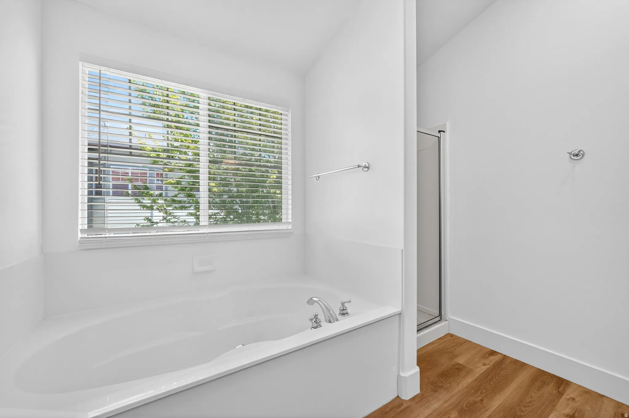 Soaking tub with chrome fixtures, large window with blinds, and wood-finish flooring