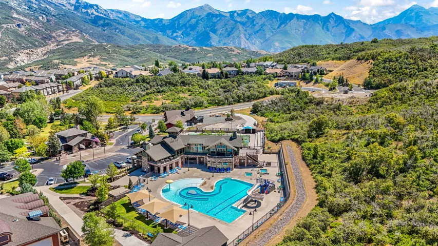 Community clubhouse featuring a multi-tiered outdoor swimming pool, adjacent lounge areas with shade structures, and a spacious parking lot