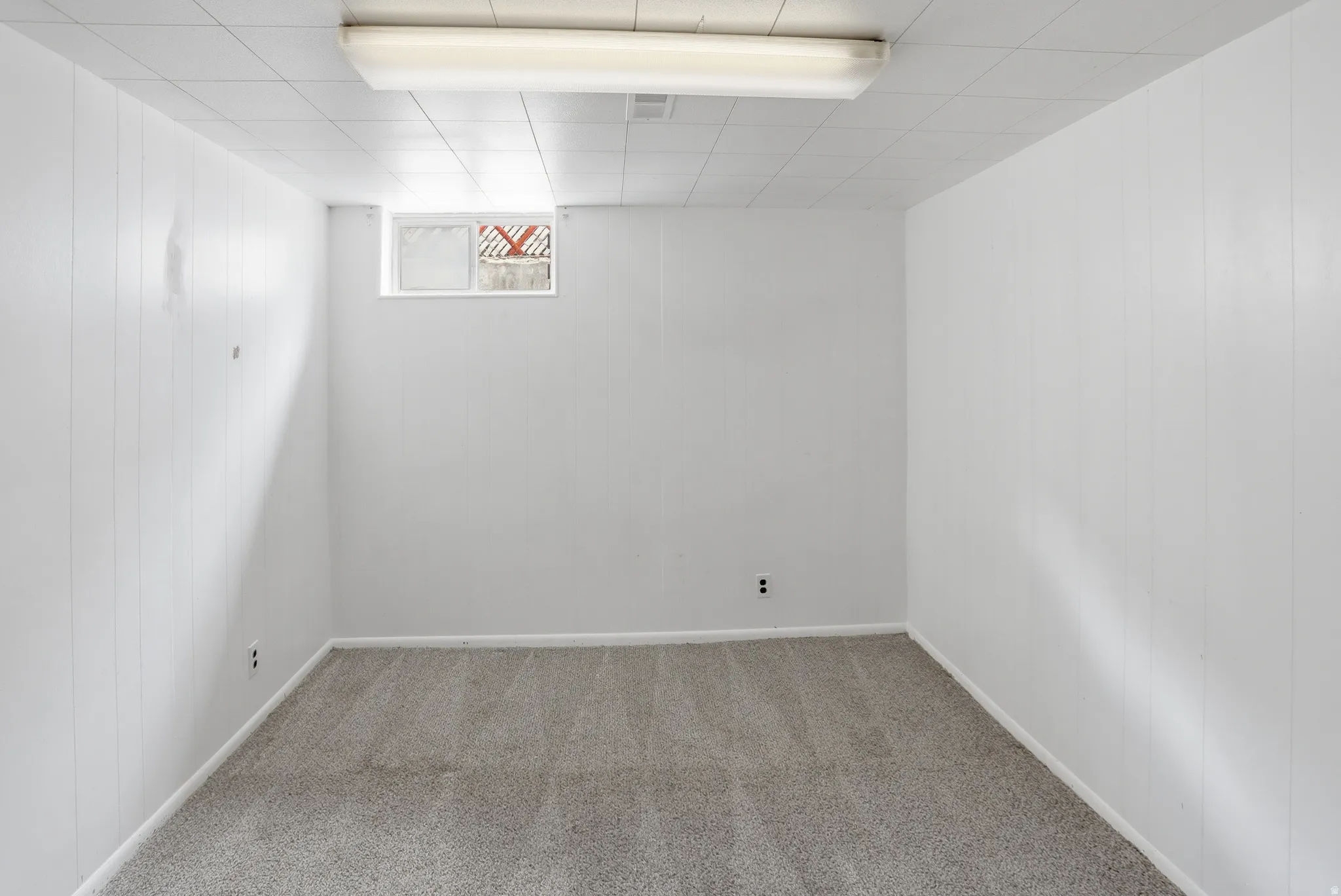 Spacious room featuring light gray carpeting, crisp white paneled walls, a drop ceiling with integrated lighting, and a single window