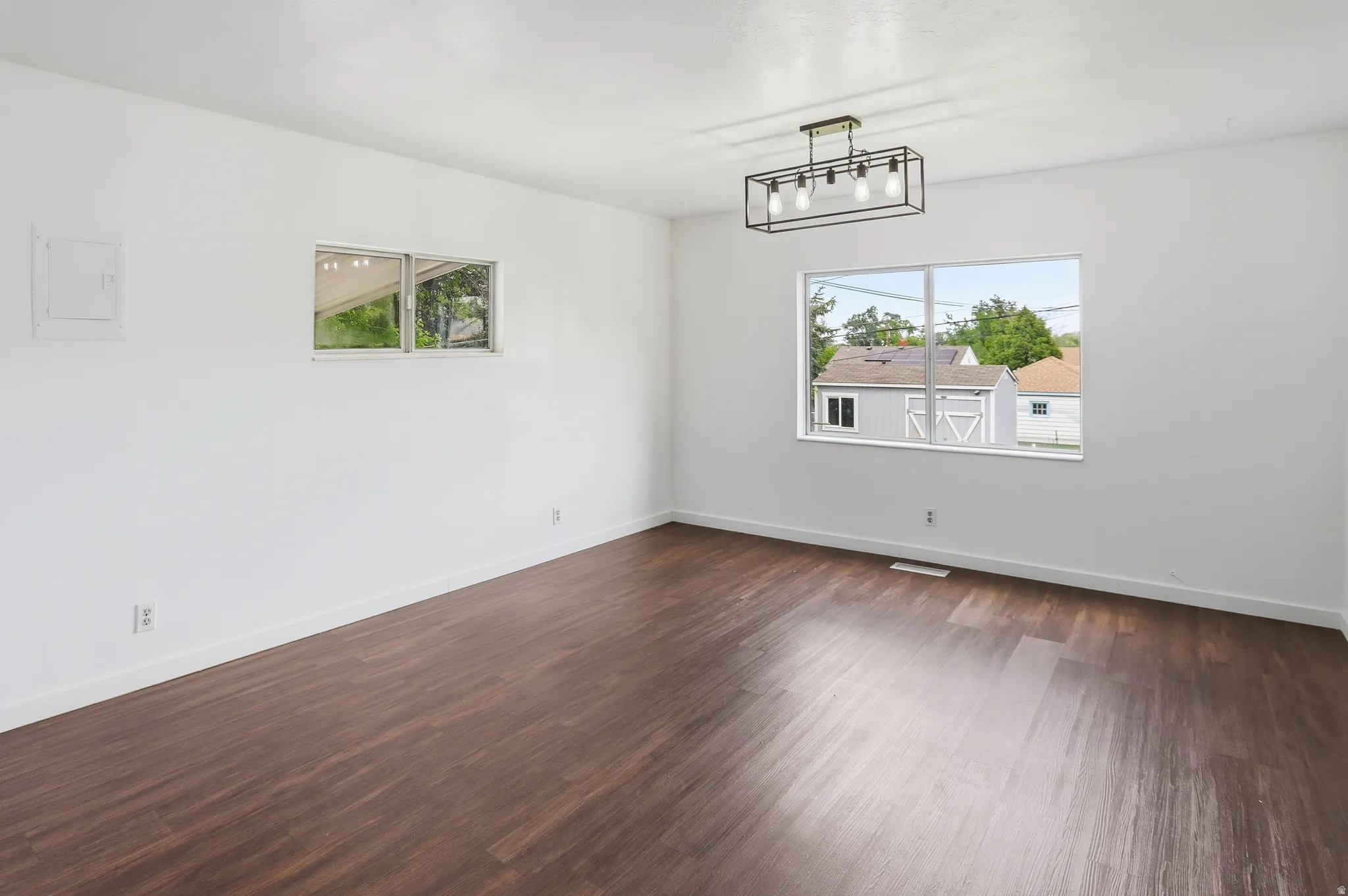 Spacious room featuring dark wood-finish flooring, crisp white walls, and a contemporary linear chandelier with exposed bulbs