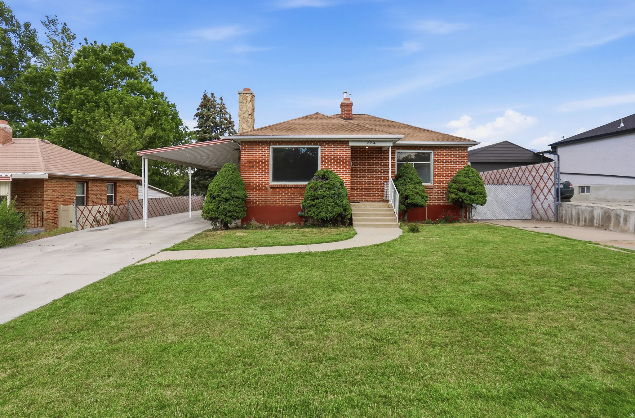 Brick exterior residence featuring a front lawn, paved driveway, covered carport, and mature shrubs