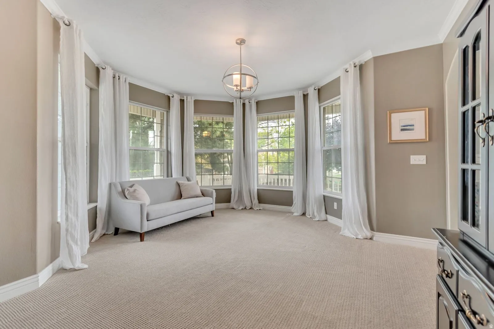 Curved wall of multiple windows, neutral tone wall paint, crown molding, light-colored textured carpet, and a modern spherical chandelier