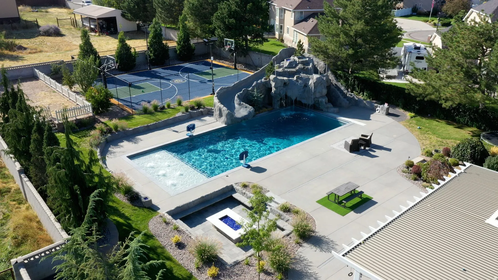 Expansive backyard oasis featuring a concrete patio, a swimming pool with a rock slide feature, a dedicated sports court, a fire pit area, and mature landscaping
