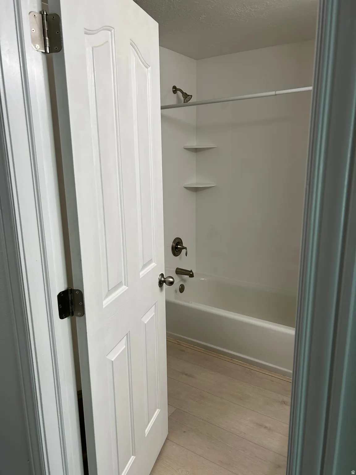 White paneled interior door opening to a bathroom featuring a shower-tub combination with brass fixtures, integrated corner shelving, and wood-finish flooring