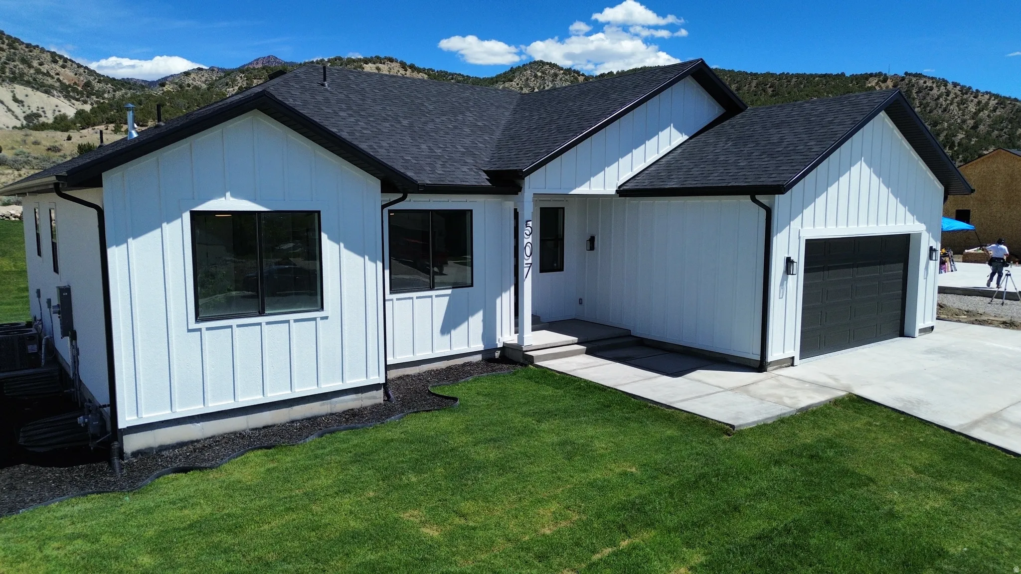 Modern farmhouse exterior featuring white vertical siding, black-framed windows, a dark asphalt shingle roof, an attached garage with a black door, and a manicured front lawn