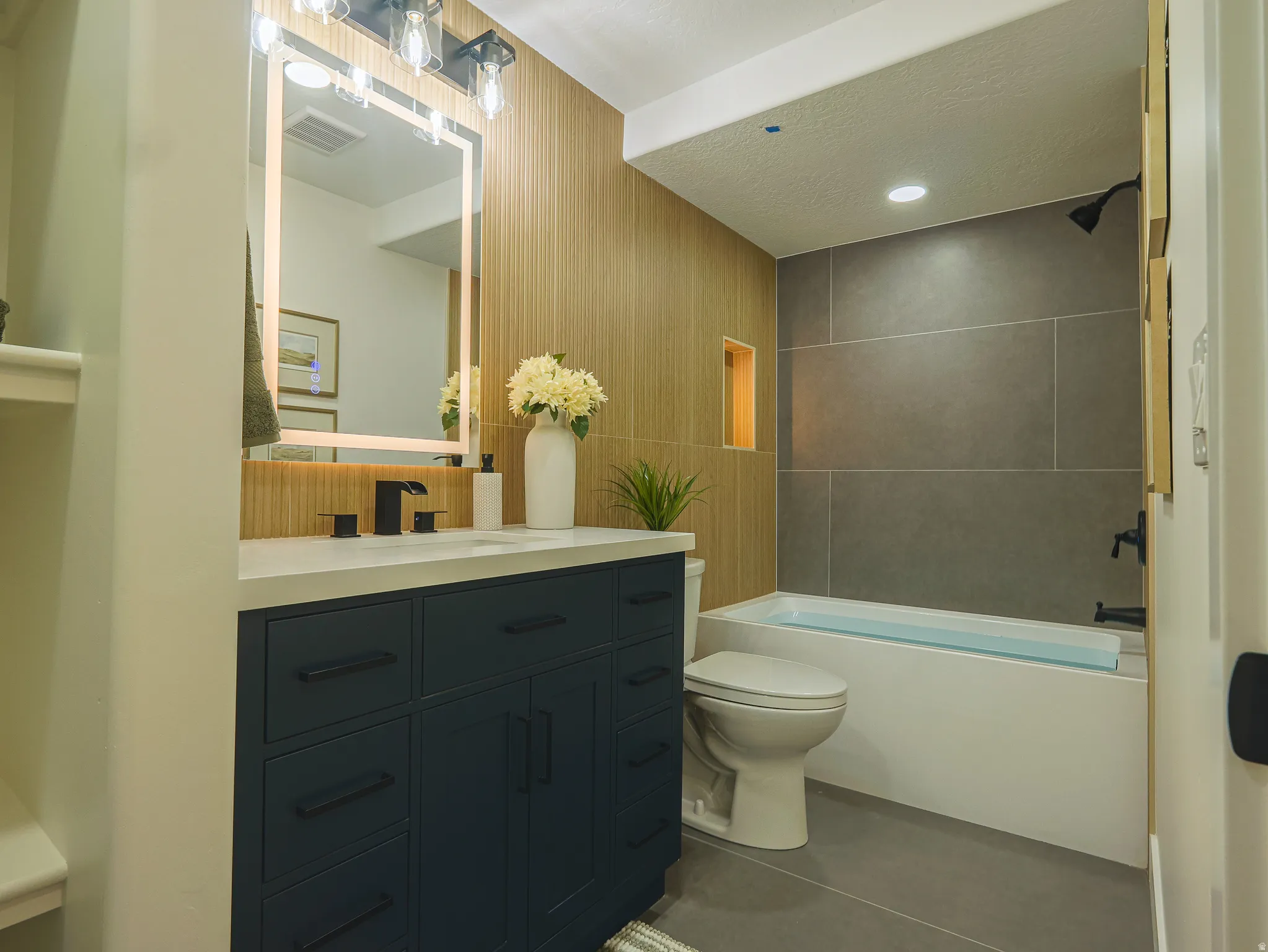 Modern bathroom featuring a dark blue vanity with a white countertop, an illuminated rectangular mirror, and a bathtub with large format gray wall tiling