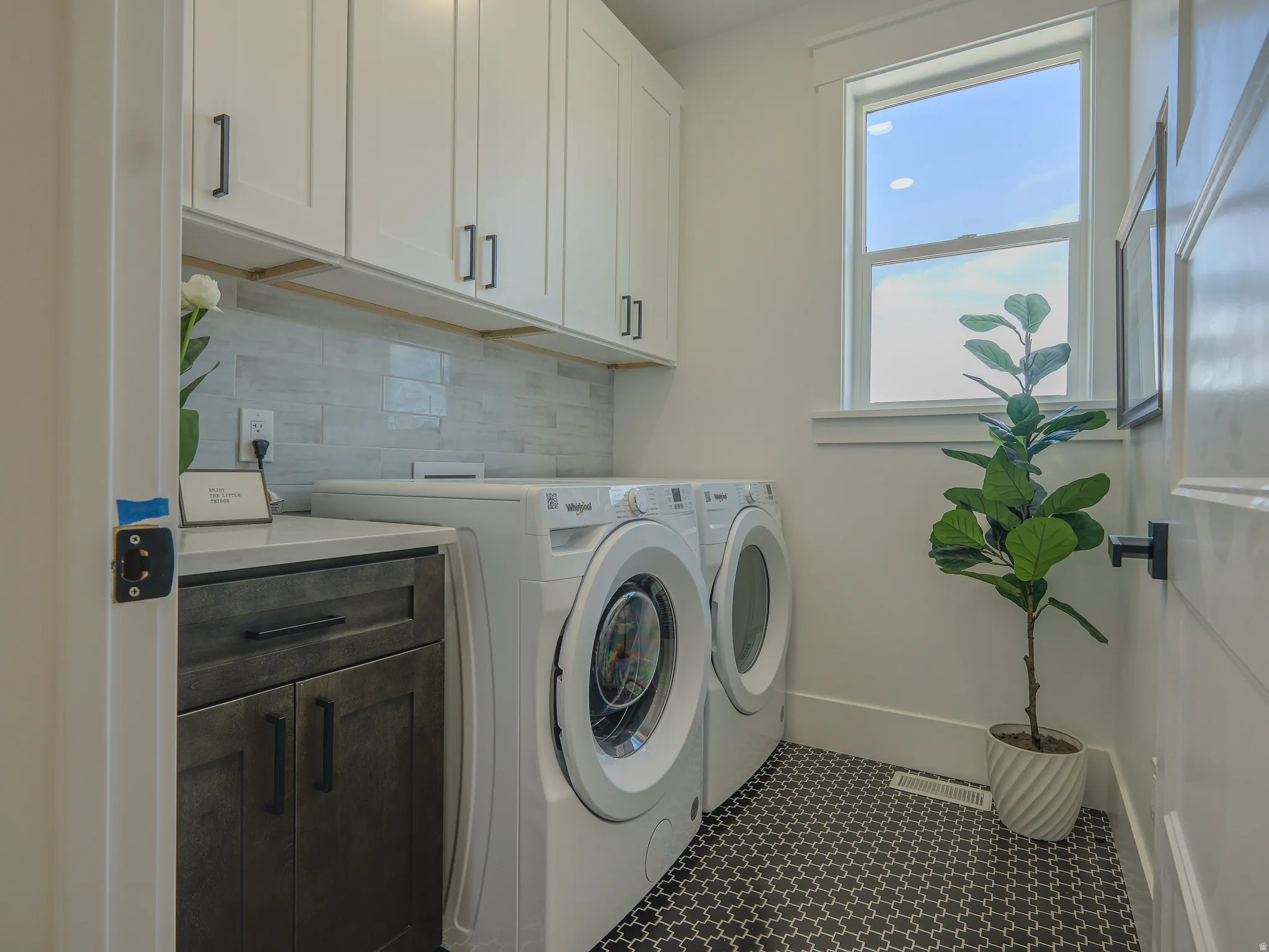 Laundry room featuring a single window, white upper cabinetry with black hardware, a dark wood-finish base cabinet with a light-toned countertop, a light gray subway tile backsplash, and dark patterned floor tiles