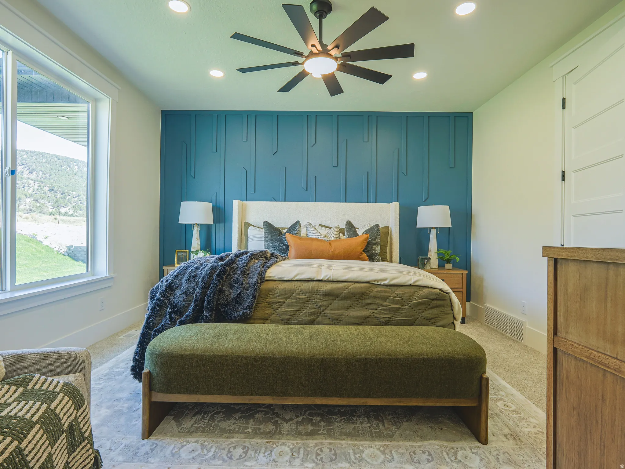 Accent wall featuring geometric paneling, large window with outdoor landscape, recessed ceiling lighting, ceiling fan with light, and light-tone carpeting
