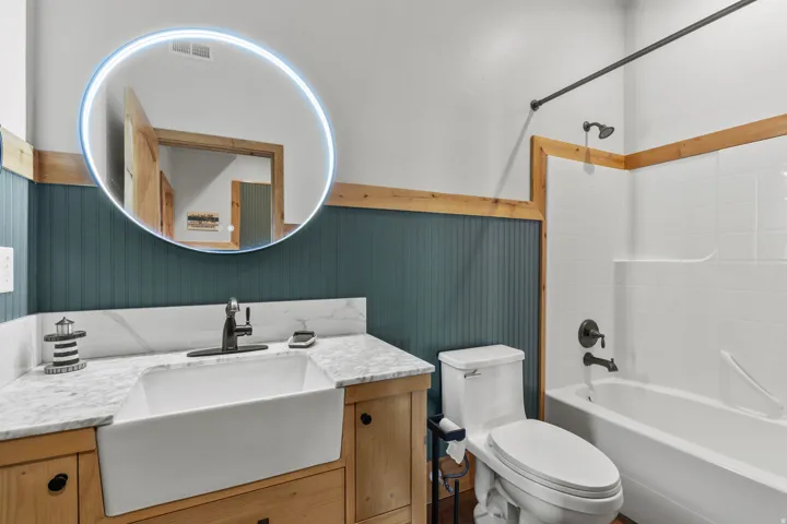 Full Bathroom featuring a farmhouse-style sink, wood-finish vanity, marble-patterned countertop, and an illuminated circular mirror
