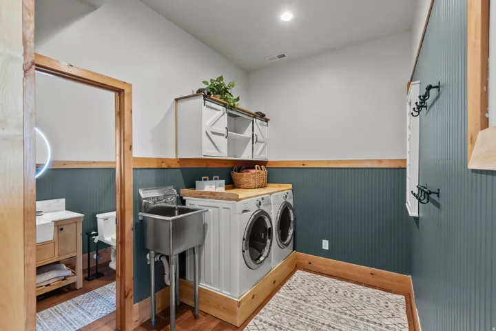Laundry room featuring a stainless steel utility sink, front-loading washer and dryer, wood-finish countertop, white upper cabinetry with barn door style fronts, and teal wainscoting