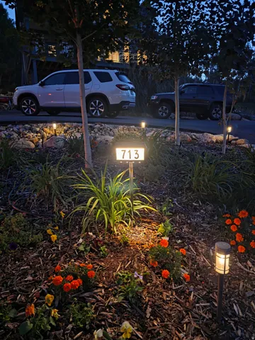 LOTS OF PARKING IN PRIVATE CIRCULAR DRIVE WAY: Illuminated address marker within a landscaped garden bed featuring varied foliage, red and yellow flowering plants, and decorative mulch