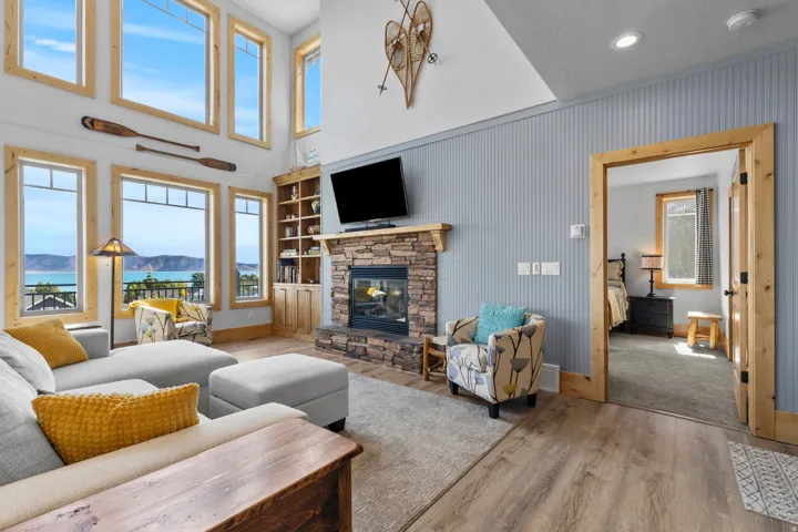 Spacious living area featuring expansive windows with water scenery, a stoned-stacked, gas fireplace with a wood mantle, wood-finish trim, a built-in wooden bookcase, and a wood framed doorway, all in the matching materials and quality.