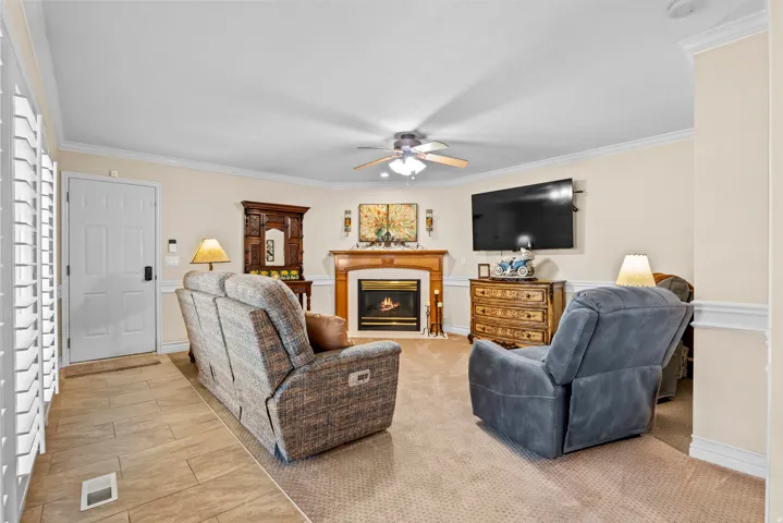 Living area featuring a fireplace with a wood-finish mantel and hearth, crown molding, a ceiling fan, and mixed flooring with tile at the entry and carpet throughout the main space