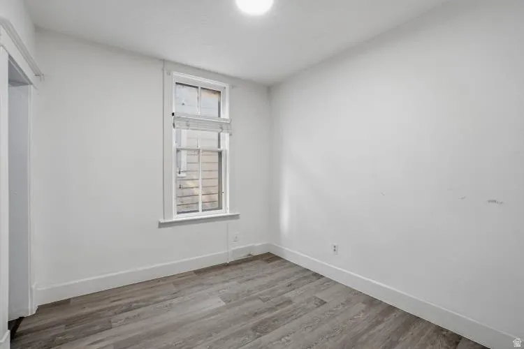 Room featuring wood-finish flooring, white baseboards, a single window with blinds, and white walls