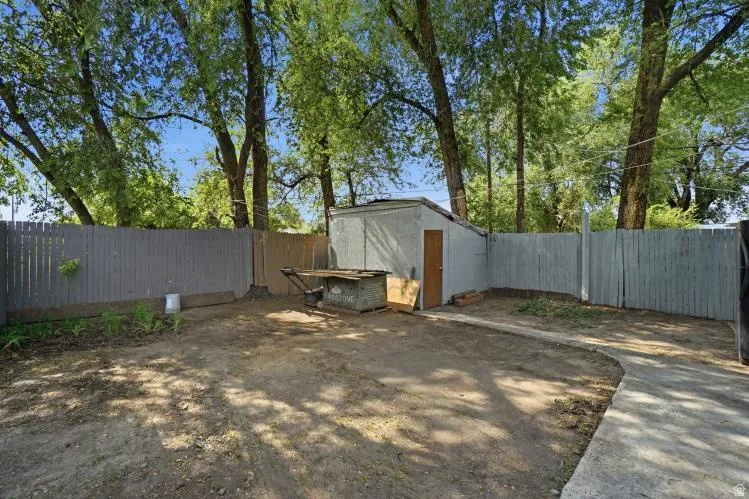 Expansive backyard featuring a concrete utility building with a wooden door, perimeter fencing, mature trees, and a concrete pathway