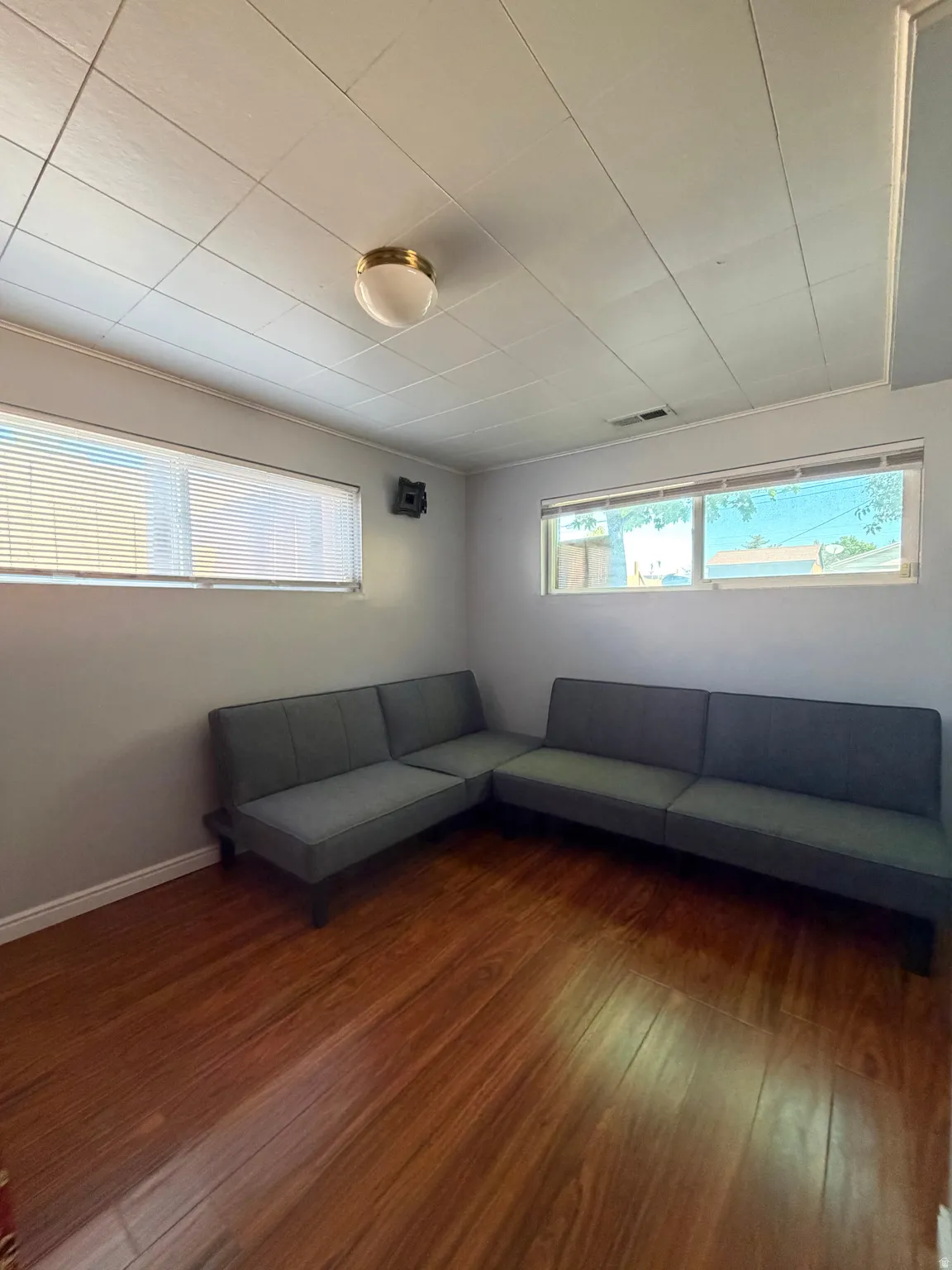 Room with laminate wood-finish flooring, two windows with blinds, and a paneled ceiling with a flush-mount light fixture