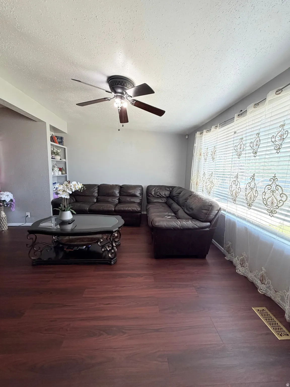 Living area featuring wood-finish flooring, a ceiling fan with integrated lighting, and a large window with blinds and sheer curtains