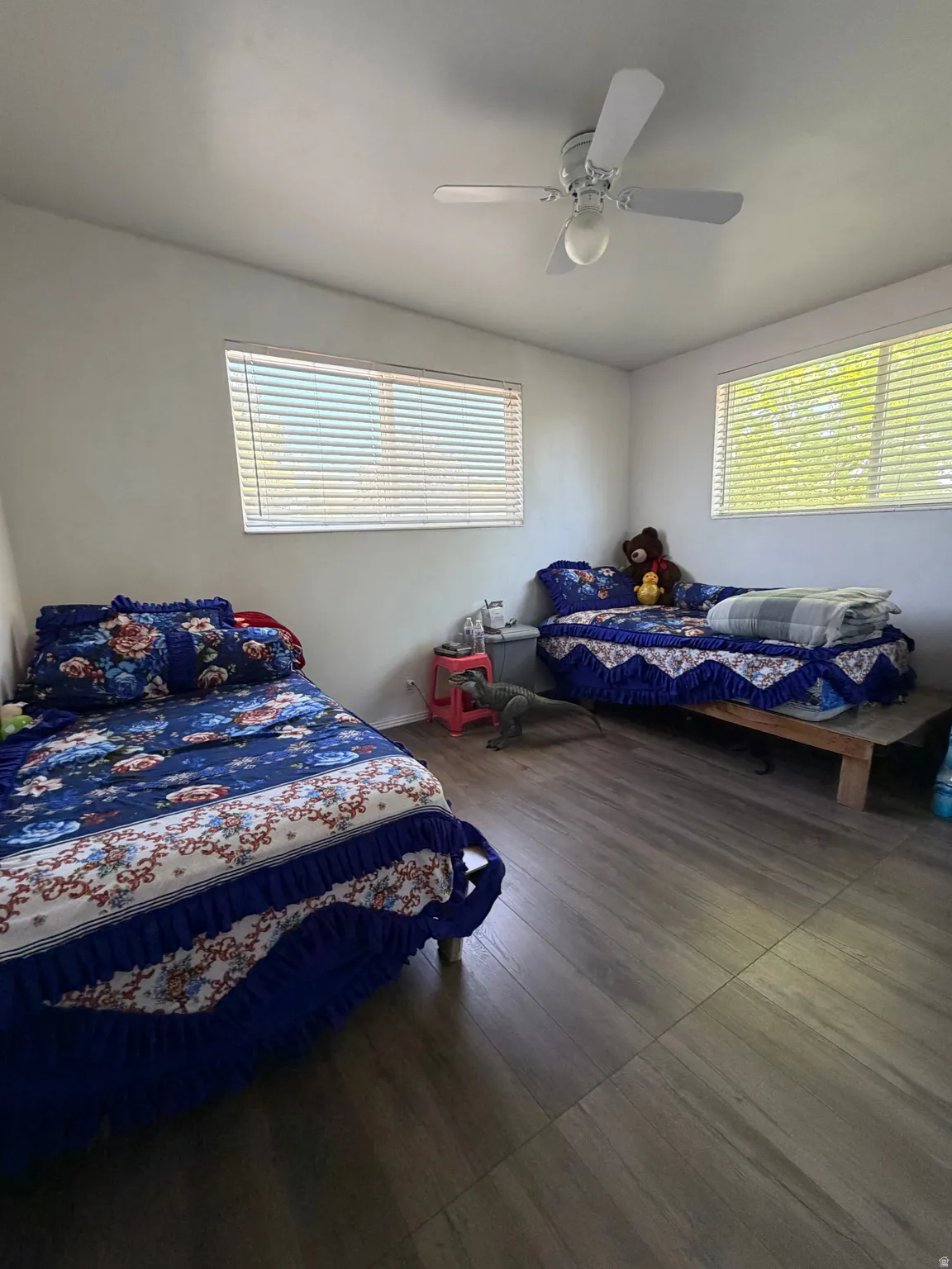 Room featuring laminate wood-finish flooring, two windows with blinds, a ceiling fan with integrated lighting, and white painted walls