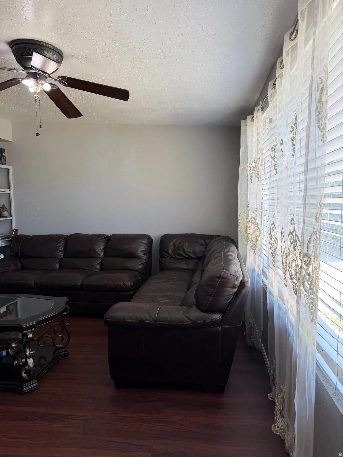 Spacious living area featuring wood-finish flooring, a ceiling fan with light fixture, and window blinds with sheer curtains