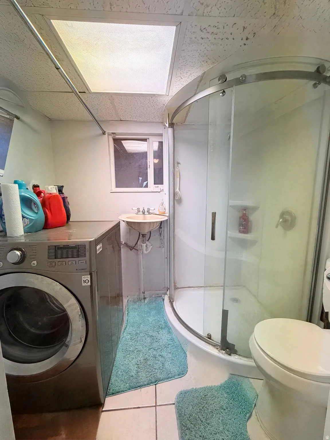 Compact bathroom featuring a curved glass-enclosed shower, a wall-mounted sink, a toilet, and tiled flooring- Laundry room