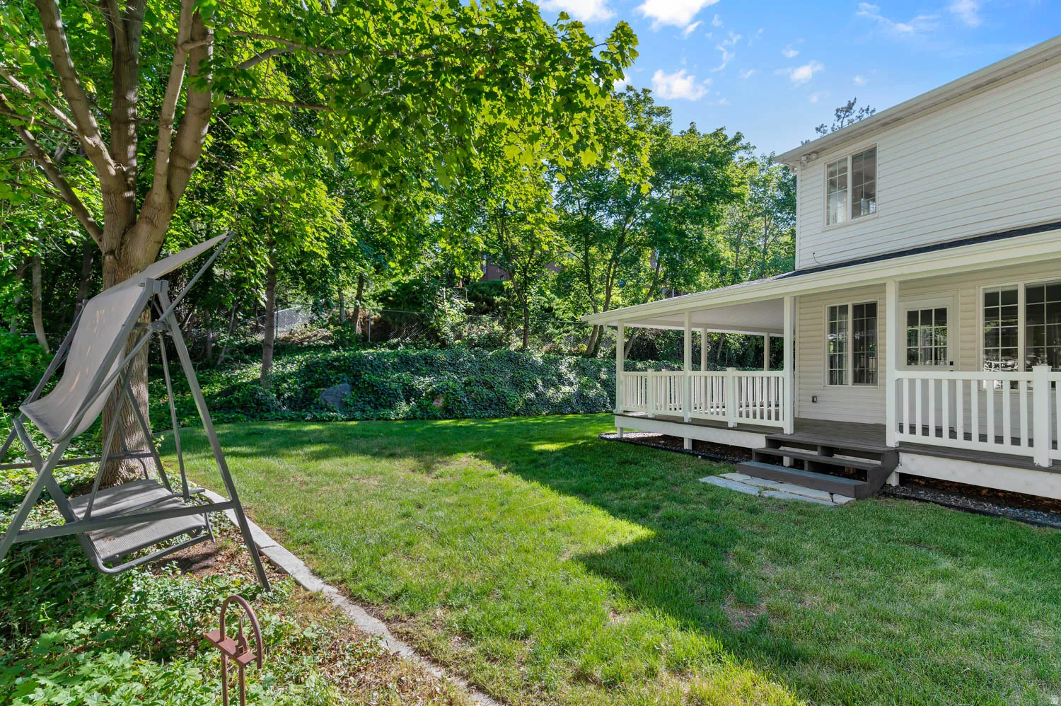 Expansive backyard featuring a covered porch with white railings, a manicured lawn, mature trees, and a two-step entry