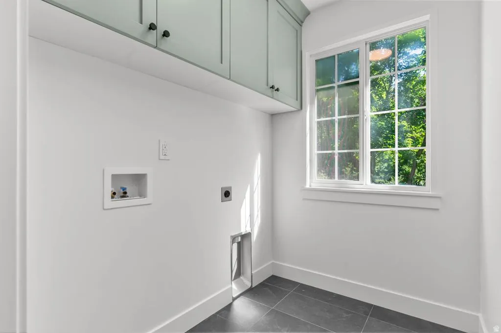Laundry room featuring light green upper cabinetry with dark hardware, a large window with gridded panes, dark tile flooring, and white baseboards